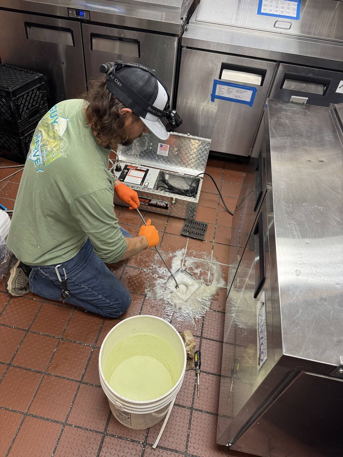 Plumber kneeling on a red-tile commercial kitchen floor, feeding a drain machine cable into a floor drain beside stainless steel coolers