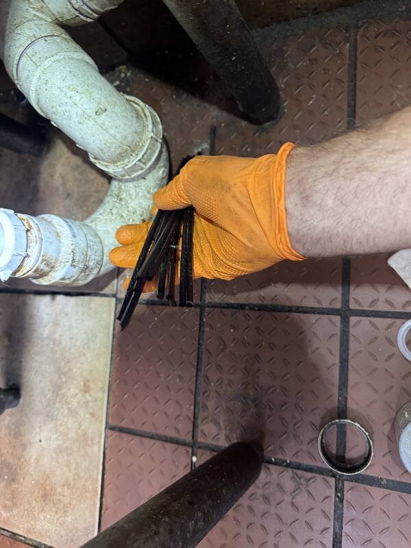 Orange-gloved hand holding black debris strips next to an opened white PVC drain line on a tile floor