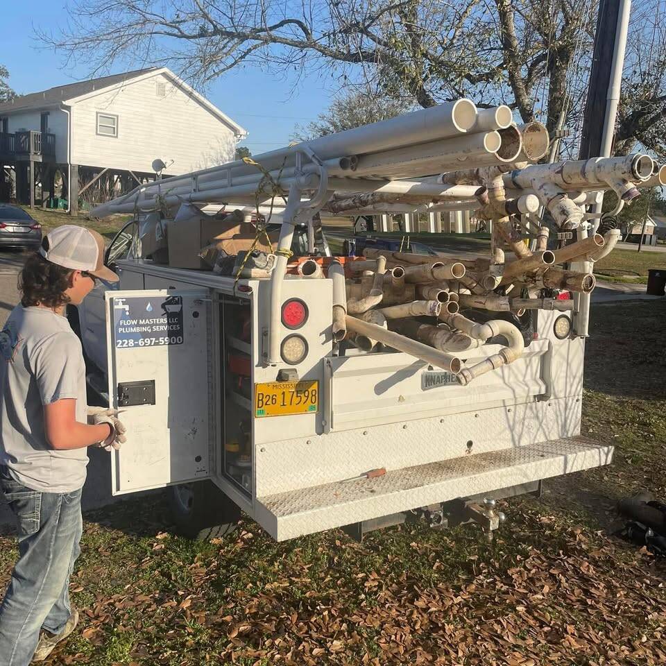 White utility-body service truck with a rack stacked high with used PVC pipe and fittings, parked under oak trees