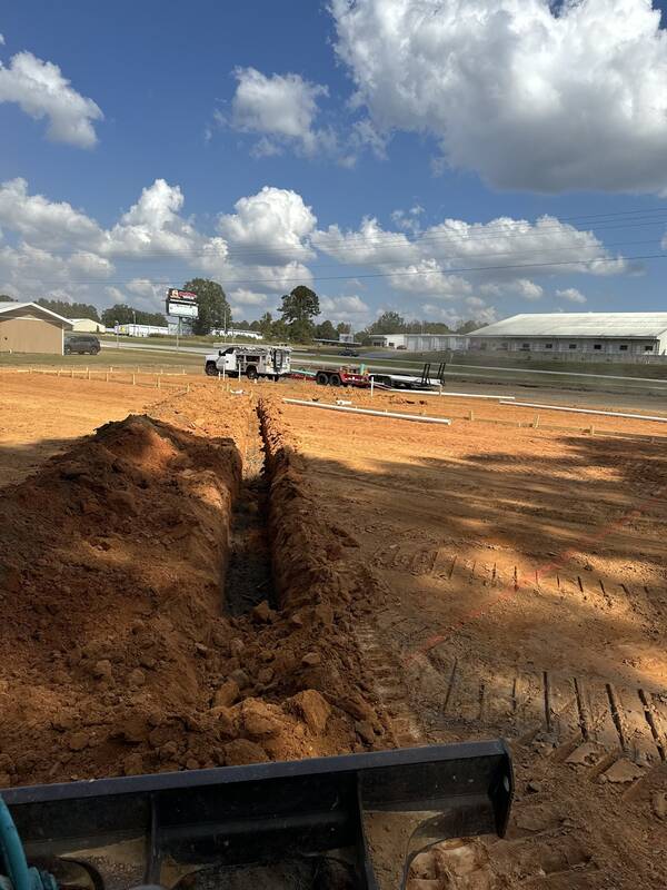 Long trench dug in red clay across a cleared lot, seen over the edge of a loader bucket
